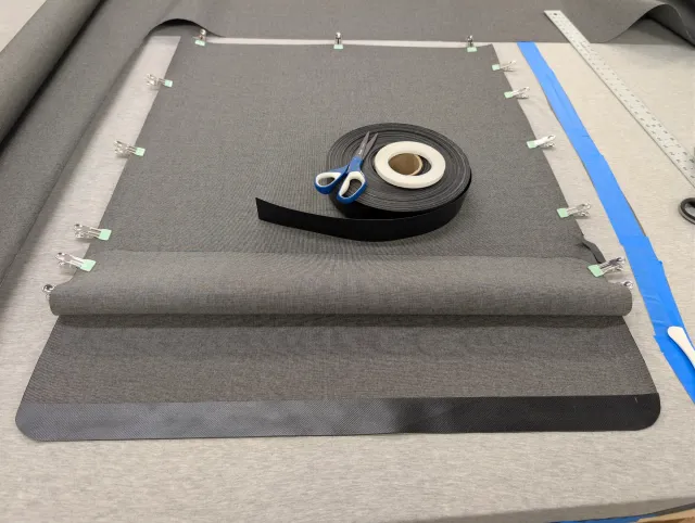 A curtain panel clipped to the bench, black edge banding folded along its lower edge, a roll of banding and scissors on top.