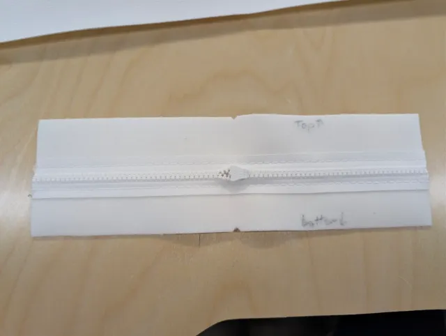 A white zipper sewn into a strip of fabric, marked top and bottom in pencil.