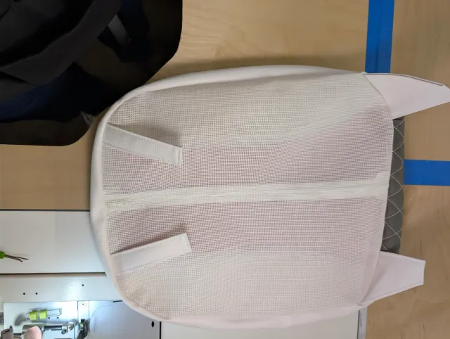 The back of a seat cushion in white mesh with a centre zipper and two straps, tabs at the corners.