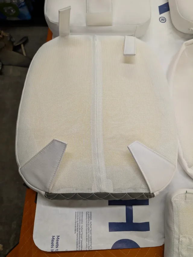 The mesh back of a seat bottom with a centre zipper and two corner tabs, charcoal edge along the bottom.