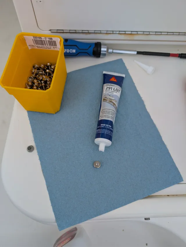 A tube of Sikaflex 291 LOT, a box of snap fastener screws and a screwdriver on a blue pad aboard the boat.
