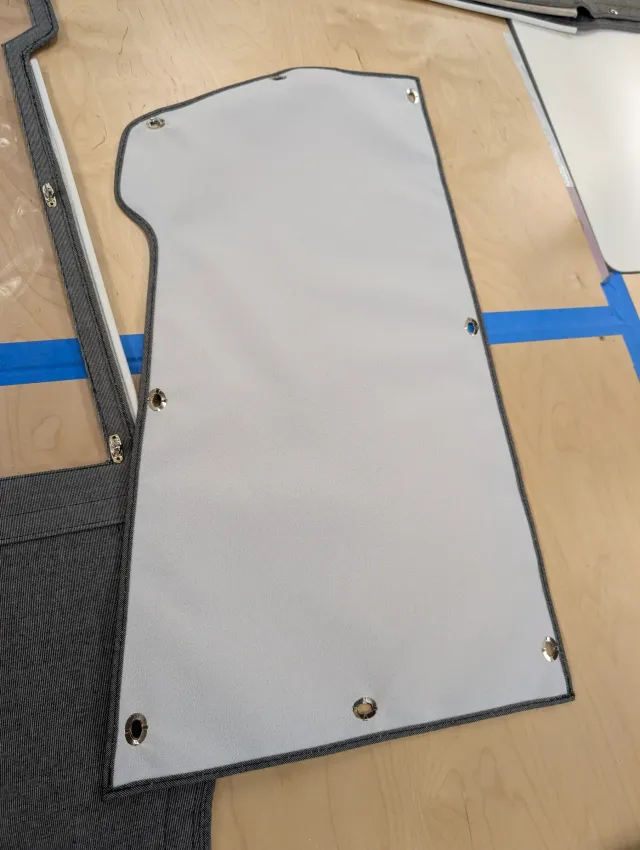 A pale grey panel with metal eyelets around its edges and grey edge binding, on the bench beside a clear-windowed curtain.