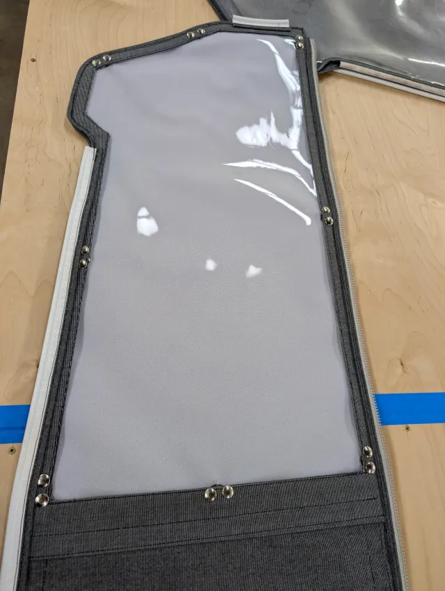 A curtain section with a clear vinyl window, pale grey fabric behind it and nickel snaps around the edge.