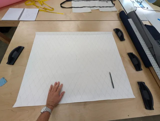 Vinyl fabric on a plywood bench, ruled with a grid of diamonds, a hand resting on it, fabric rolls at the right.