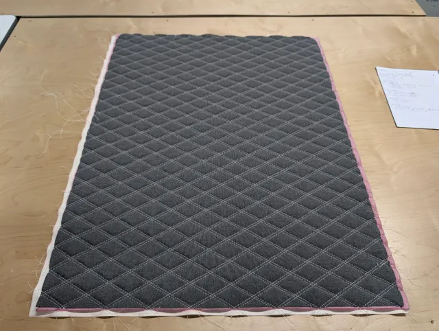 A grey fabric panel quilted in diamonds with white stitching, laid on the workbench with loose threads at the edges.
