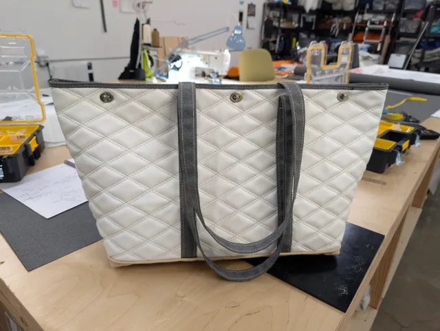 The finished white quilted tote standing on a bench, with grey straps, three twist-lock closures and a tan base.