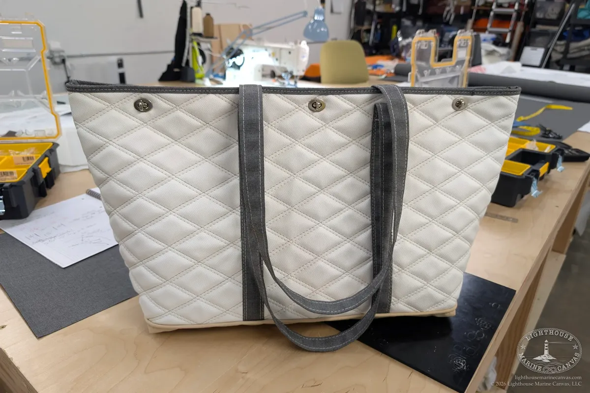 The finished white quilted tote standing on a bench, with grey straps, three twist-lock closures and a tan base.