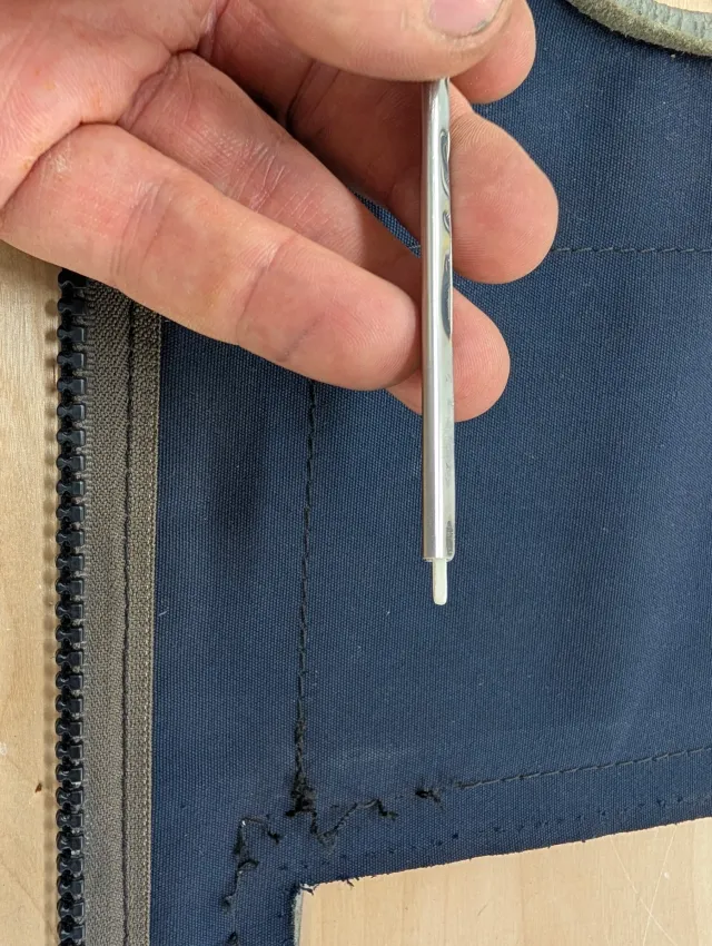 A hand holding a slim steel fabric marking tool over the old stitch line of a navy canvas panel, dark thread remnants still in the cloth beside the zipper.