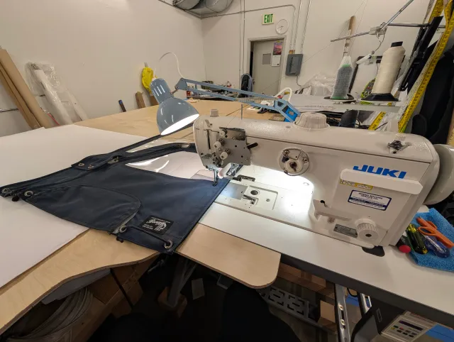 A navy enclosure panel under the needle of an industrial sewing machine on the workbench, a desk lamp lighting the work and a cone of thread behind.