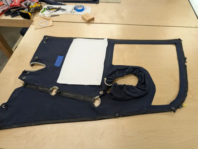 A navy enclosure panel flat on the workbench, a white protective cover laid over one window, zippers along its edges and a gathered sleeve at one corner.