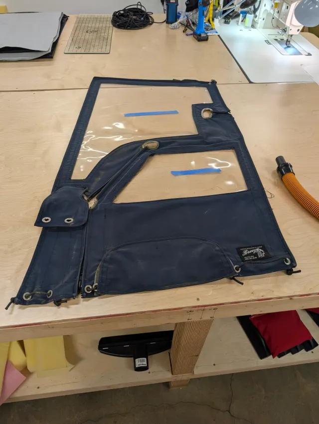 An old navy enclosure panel laid flat on the workbench, its two fogged vinyl windows marked with strips of blue tape and the zipper edges around it.