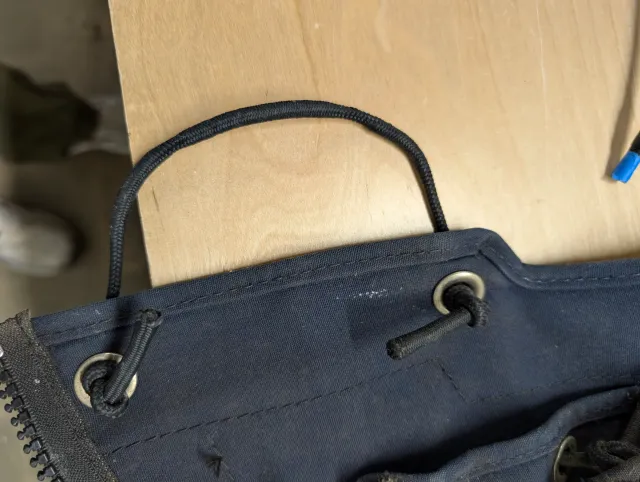 The lower edge of a navy enclosure panel on the bench, a loop of old black shock cord run through two stainless grommets and knotted on the inside.